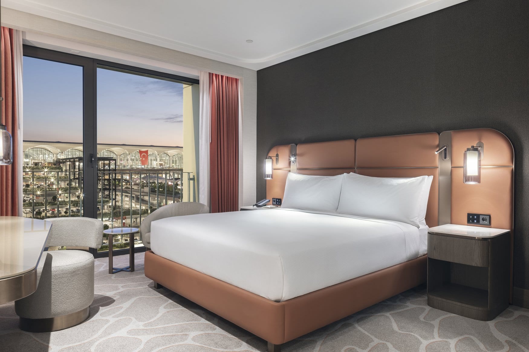 Hotelzimmer im Hilton Istanbul Airport mit Doppelbett vor einem Kopfteil aus cognacfarbenem Leder. Das bodentiefe Fenster gibt den Blick auf das beleuchtete Terminal frei.