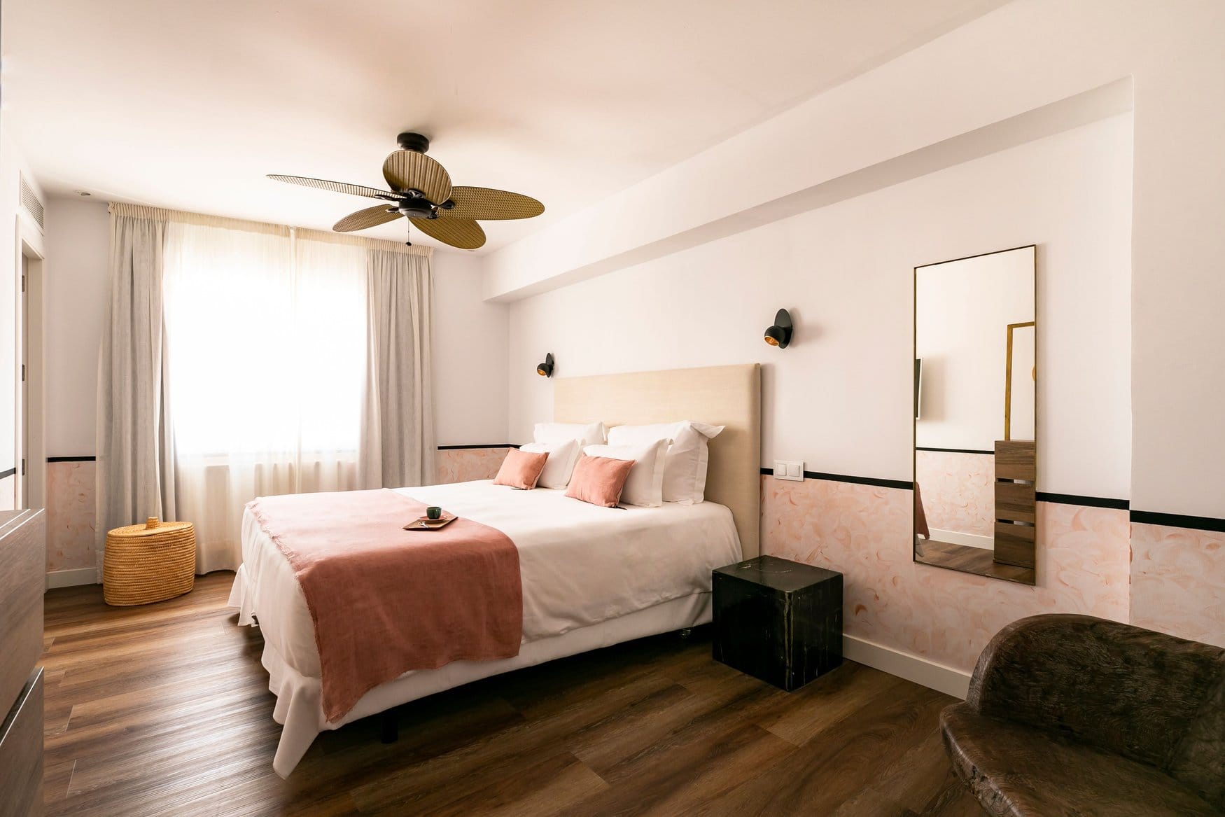 Hotelzimmer im Mikasa mit Doppelbett, Deckenventilator aus Korbgeflecht und einem Standspiegel an der Wand. Die untere Wandhälfte ist in Altrosa gestrichen, auf dem Boden liegt dunkles Holzparkett.
