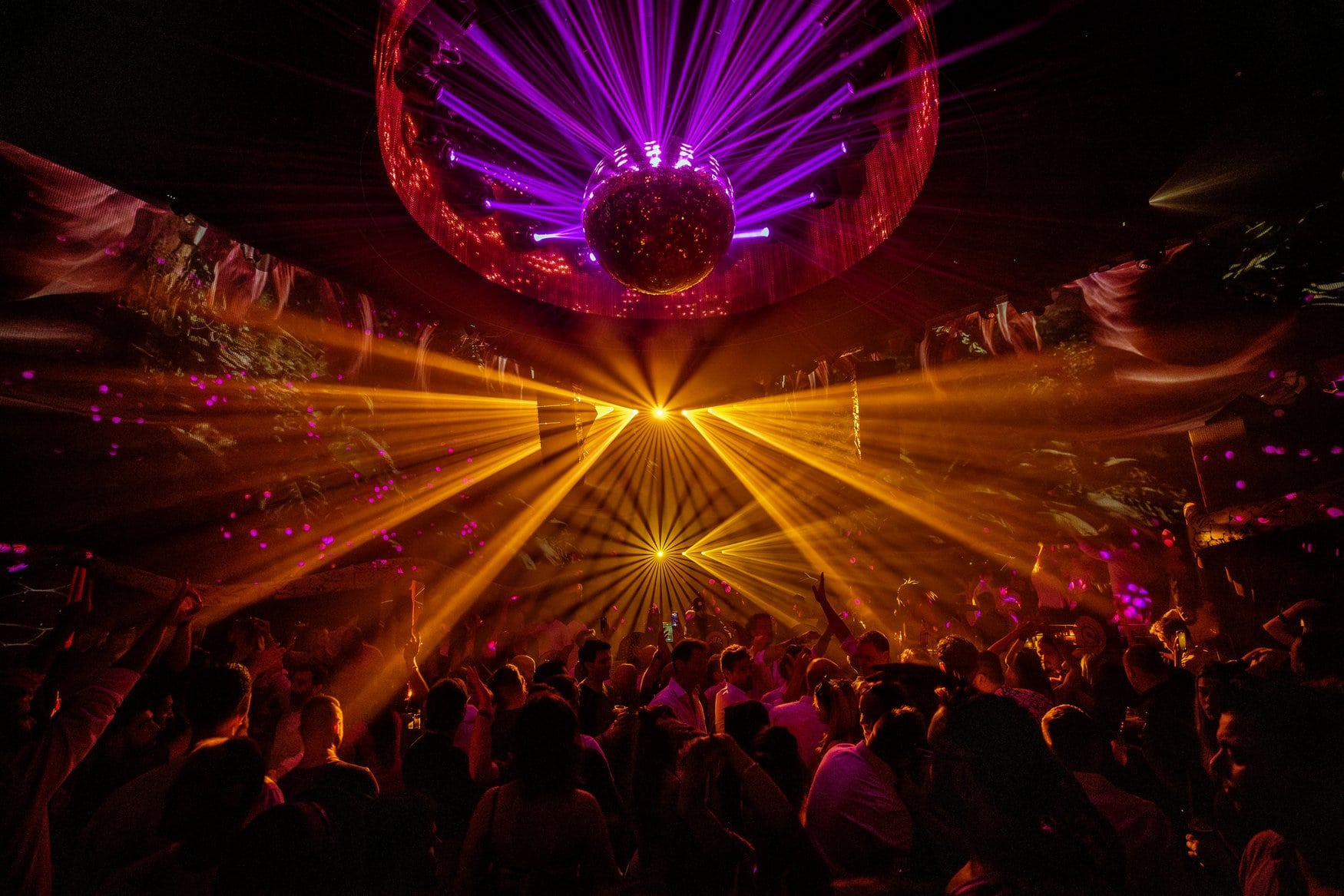 Tanzfläche im Club Chinois auf Ibiza: eine große Diskokugel unter der Decke wirft violette Lichtstrahlen über die tanzende Menge, darunter fächern gelbe Scheinwerferkegel auf.