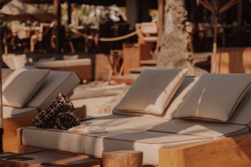 Daybed mit Kissen und Handtüchern im Sand des Strandclubs Beachhouse. Im Hintergrund stehen Sonnenschirme und Palmen.