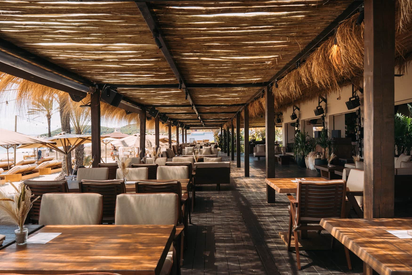 Überdachter Gastraum des Strandclubs Beachhouse auf Ibiza mit Holztischen unter einem Dach aus Rundhölzern und Schilf. Links öffnet sich der Blick auf Sonnenschirme am Strand.