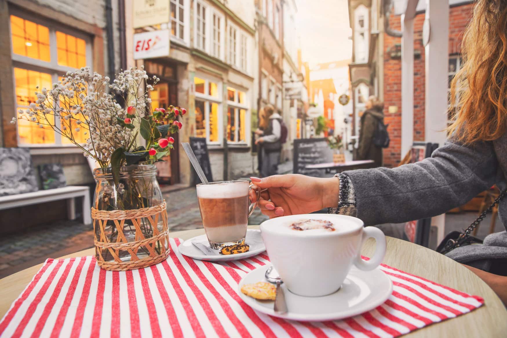 Cappuccino und Latte macchiato auf einem Tisch im Straßencafé im Schnoorviertel, dahinter beleuchtete Schaufenster und Passanten.