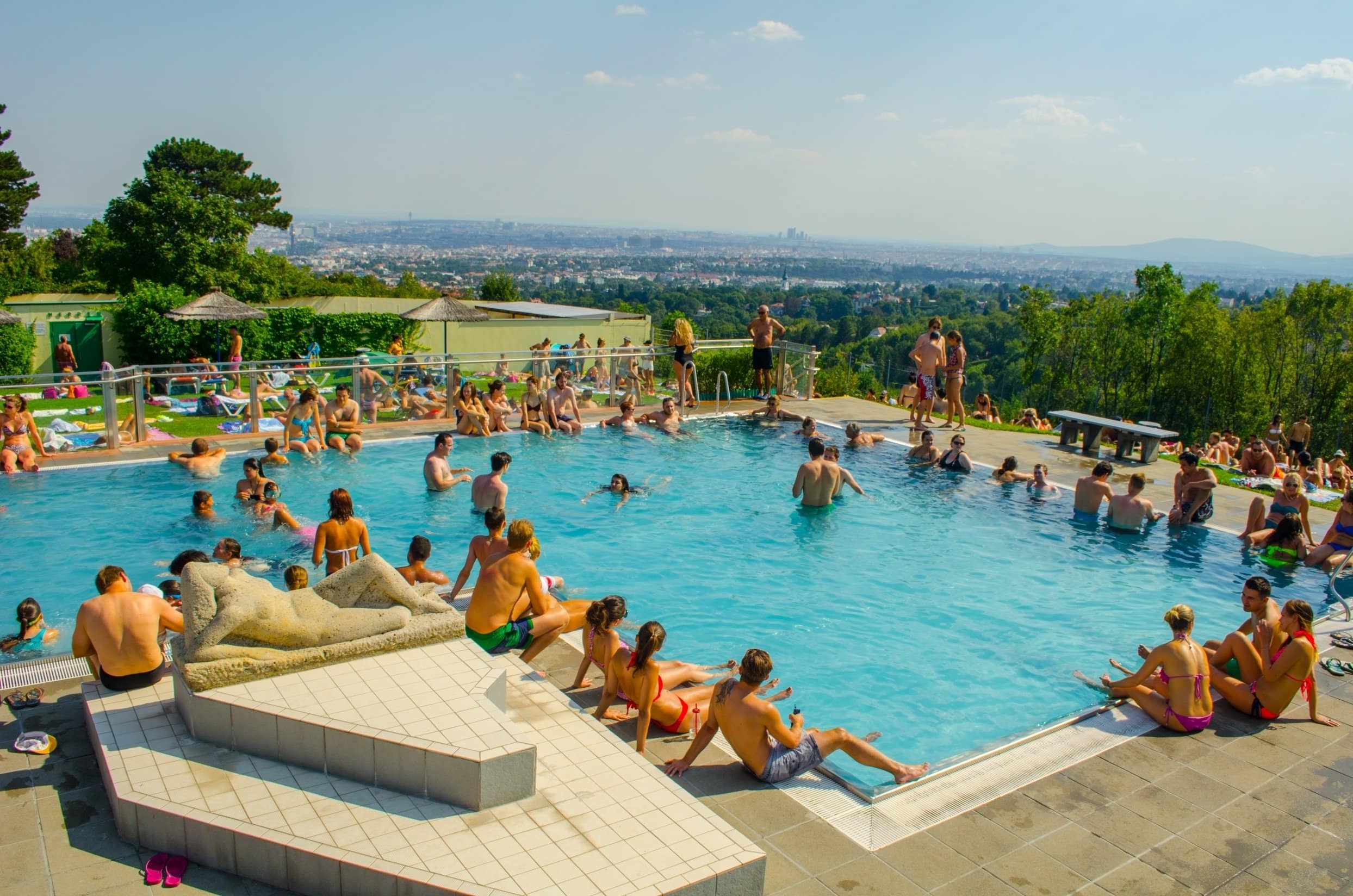 Badegäste im Becken des Krapfenwaldlbads in Wien, im Hintergrund reicht der Blick über die Stadt bis zum Horizont.