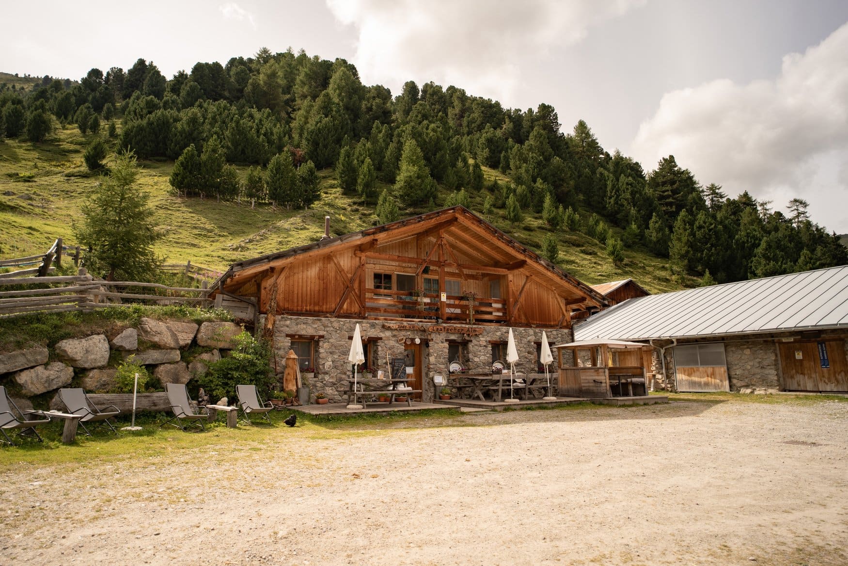Die Laatscher Alm mit Steinsockel und Holzfassade vor einem bewaldeten Hang, davor Sitzplätze und Sonnenschirme.