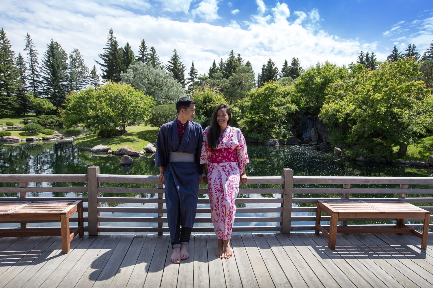 Paar in Yukata steht auf der Holzbrücke über dem Teich des Nikka Yuko Japanese Garden in Lethbridge, dahinter Kiefern und ein kleiner Wasserfall