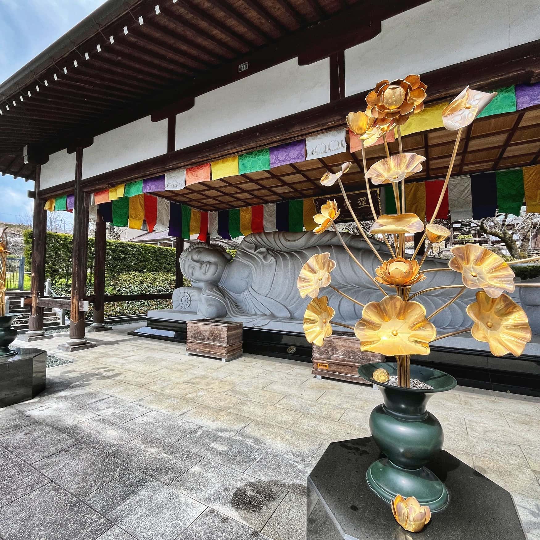 Eine liegende Buddha-Statue aus Stein unter bunten buddhistischen Bannern, davor eine goldene Lotusblumen-Skulptur, am Ishiteji-Tempel Nummer 51 des Pilgerwegs auf Shikoku.