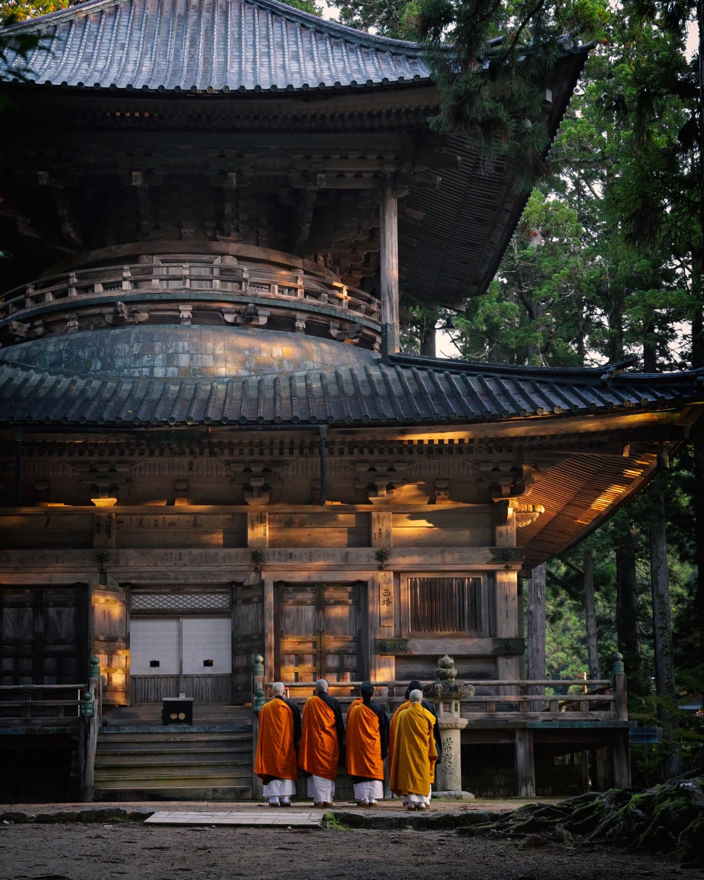 Mehrere Mönche in orangefarbenen und gelben Gewändern stehen bei Abendlicht vor einer großen historischen Tempelhalle auf dem Koyasan.