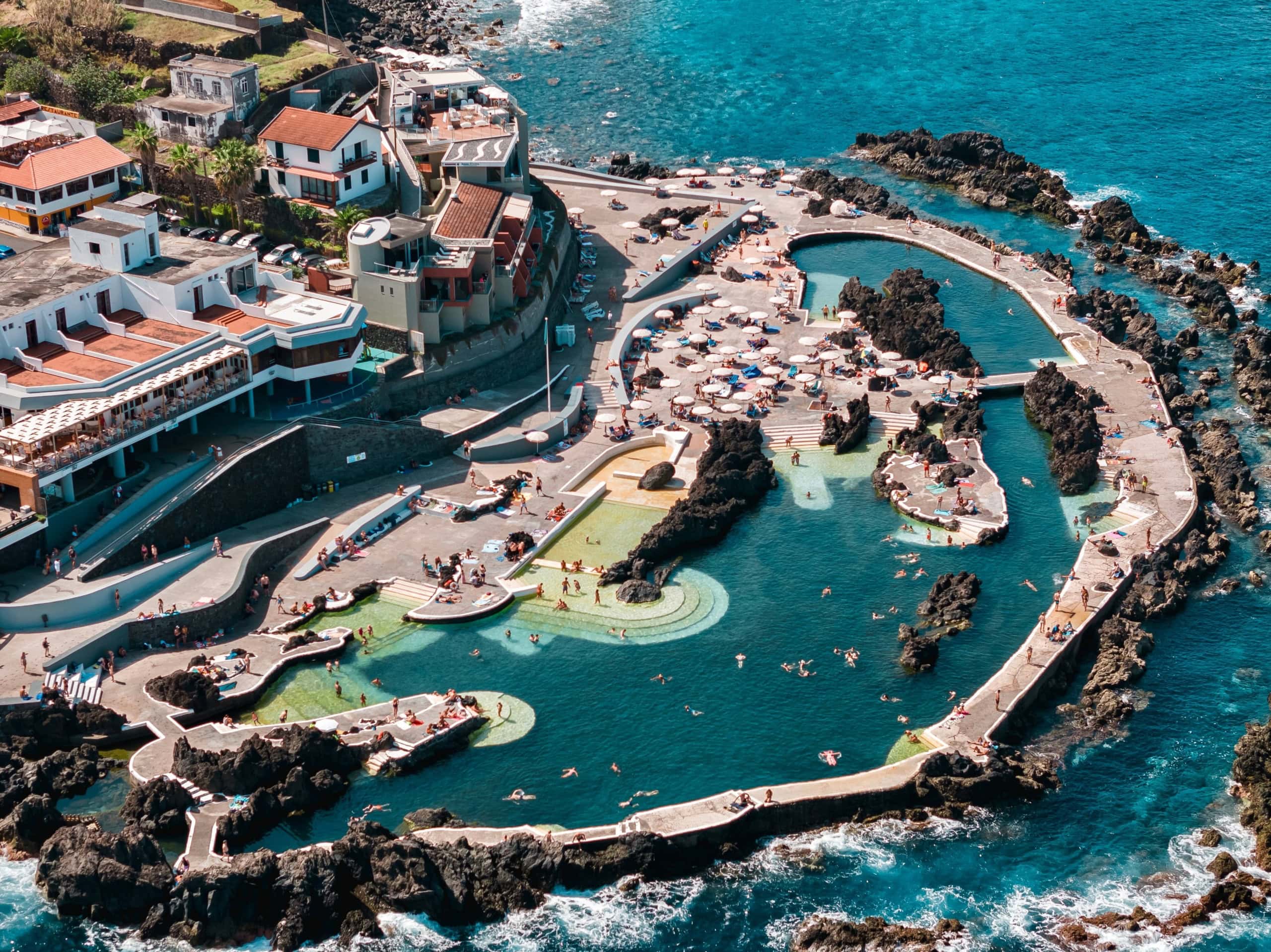 Luftaufnahme der Naturpools von Porto Moniz auf Madeira mit dunklen Vulkanfelsen und türkisfarbenem Wasser. Sonnenterrassen mit Liegestühlen umgeben die Becken.