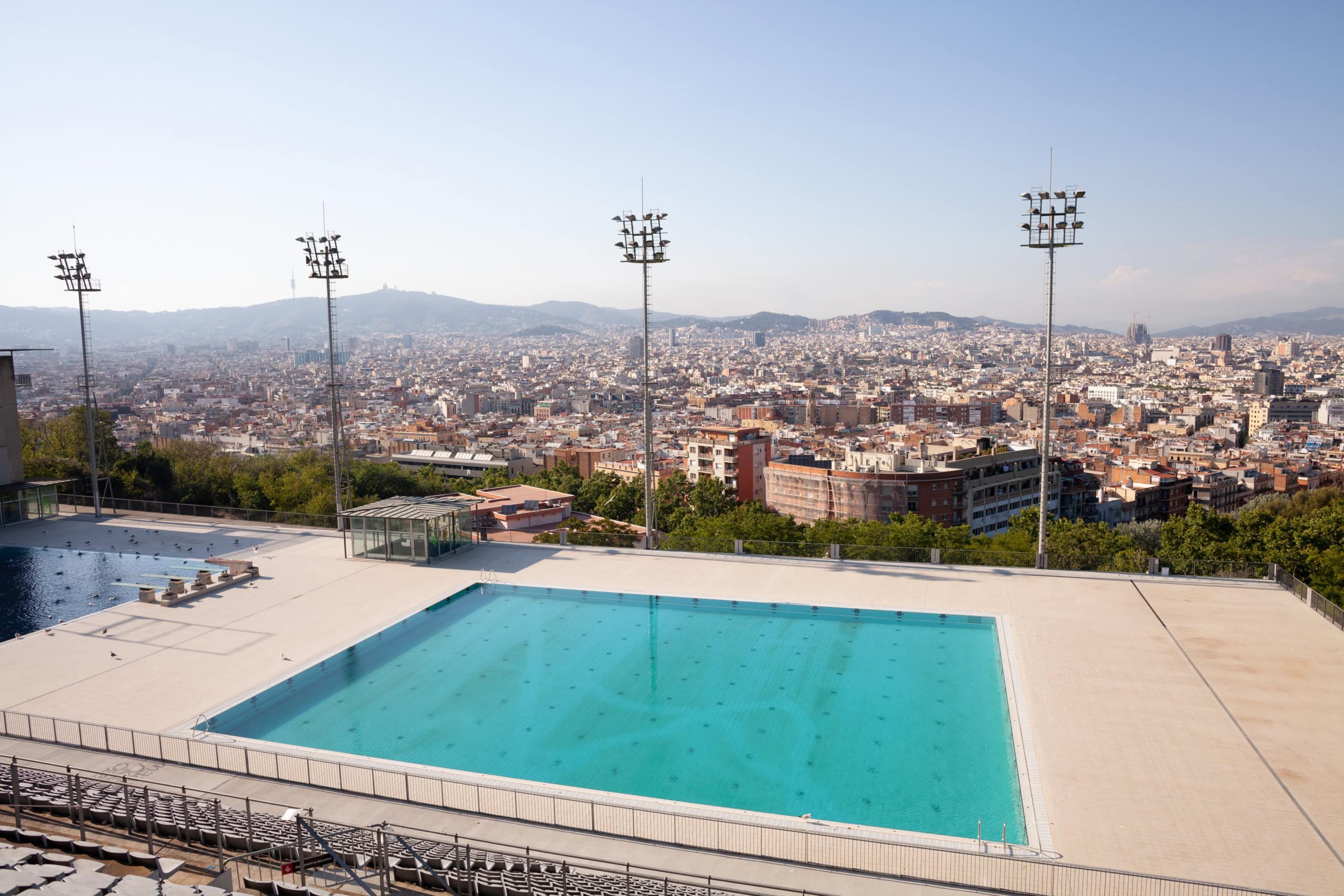 Sprungbecken der Piscina Municipal de Montjuïc in Barcelona mit Flutlichtmasten, im Hintergrund erstreckt sich die Stadt bis zum Horizont.