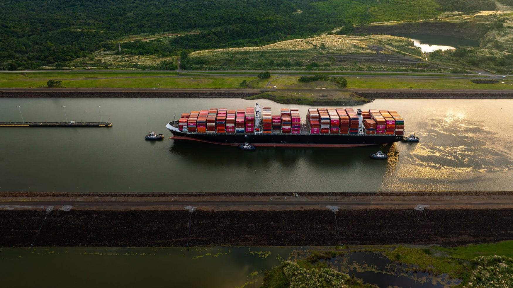 Luftaufnahme eines beladenen Containerschiffs im Panamakanal, das von vier Schleppern begleitet wird. Die Fahrrinne verläuft zwischen befestigten Böschungen und bewaldeten Hügeln.