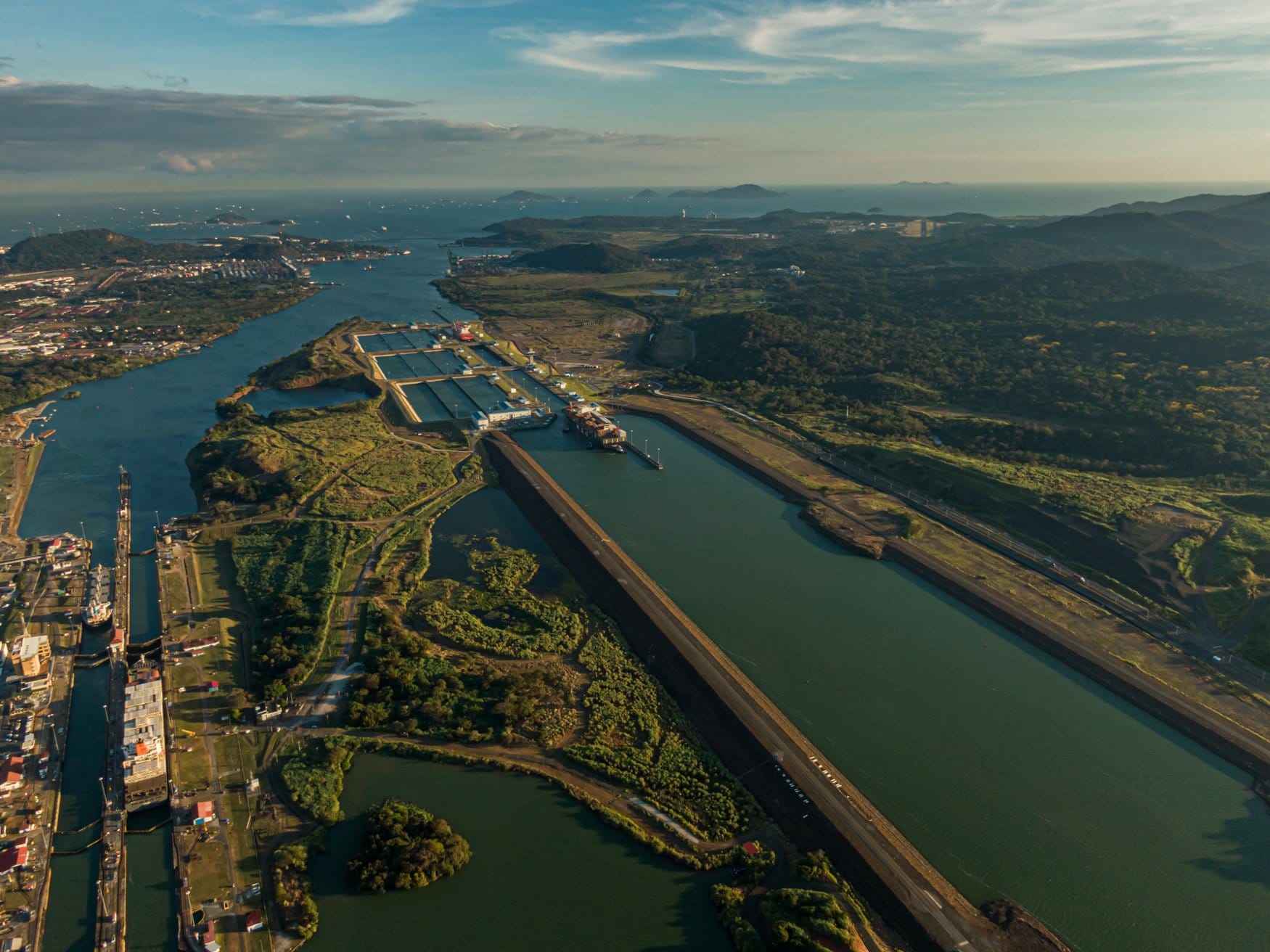Luftaufnahme der Miraflores-Schleusen am Panamakanal im Abendlicht. Im Hintergrund öffnet sich die Bucht von Panama zum Pazifik, an den Kammern liegen Containerschiffe.