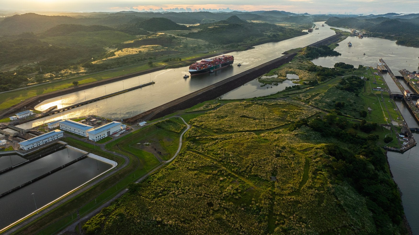 Luftaufnahme einer Schleusenanlage am Panamakanal im Morgenlicht. Ein Containerschiff wird von Schleppern durch die Kanalrinne begleitet, links liegen die Wasserspeicherbecken der Schleuse.