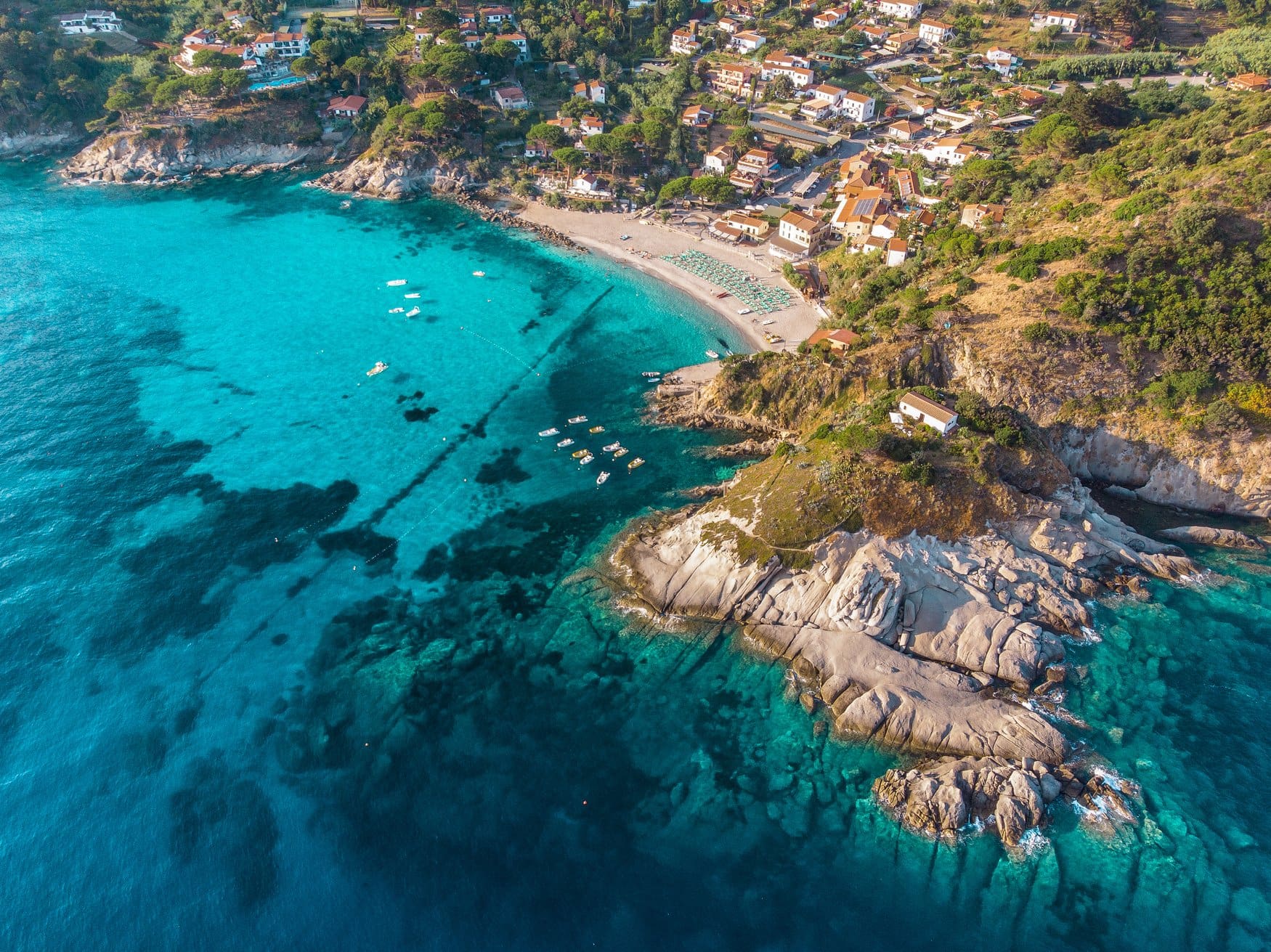 Luftaufnahme der Bucht von Sant'Andrea bei Marciana auf Elba mit türkisfarbenem Wasser, hellen Granitfelsen und einem Sandstrand mit Liegereihen.
