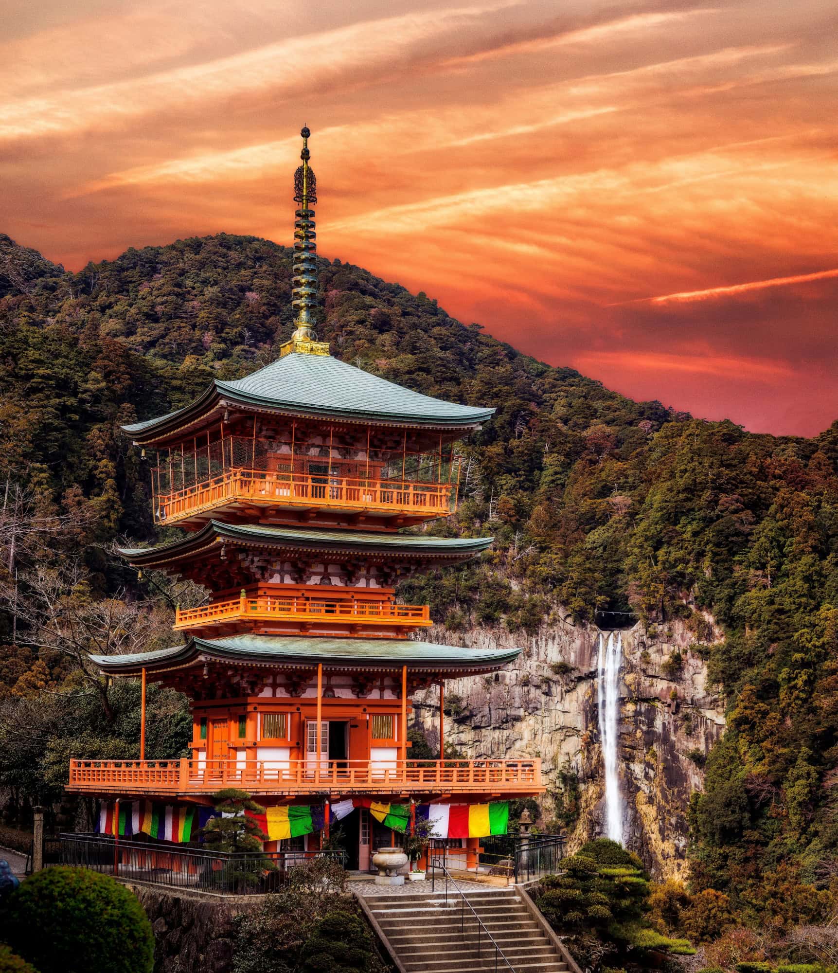 Die dreistöckige Pagode des Seiganto-ji-Tempels vor dem Nachi-Wasserfall unter einem dramatisch orange gefärbten Abendhimmel.