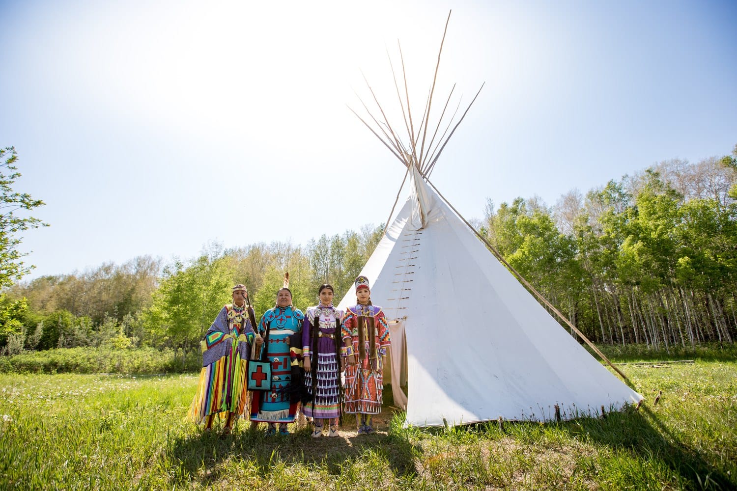 Vier Tänzerinnen und Tänzer der Siksika Nation in Powwow-Regalia stehen vor einem weißen Tipi am Waldrand im Tipi Village