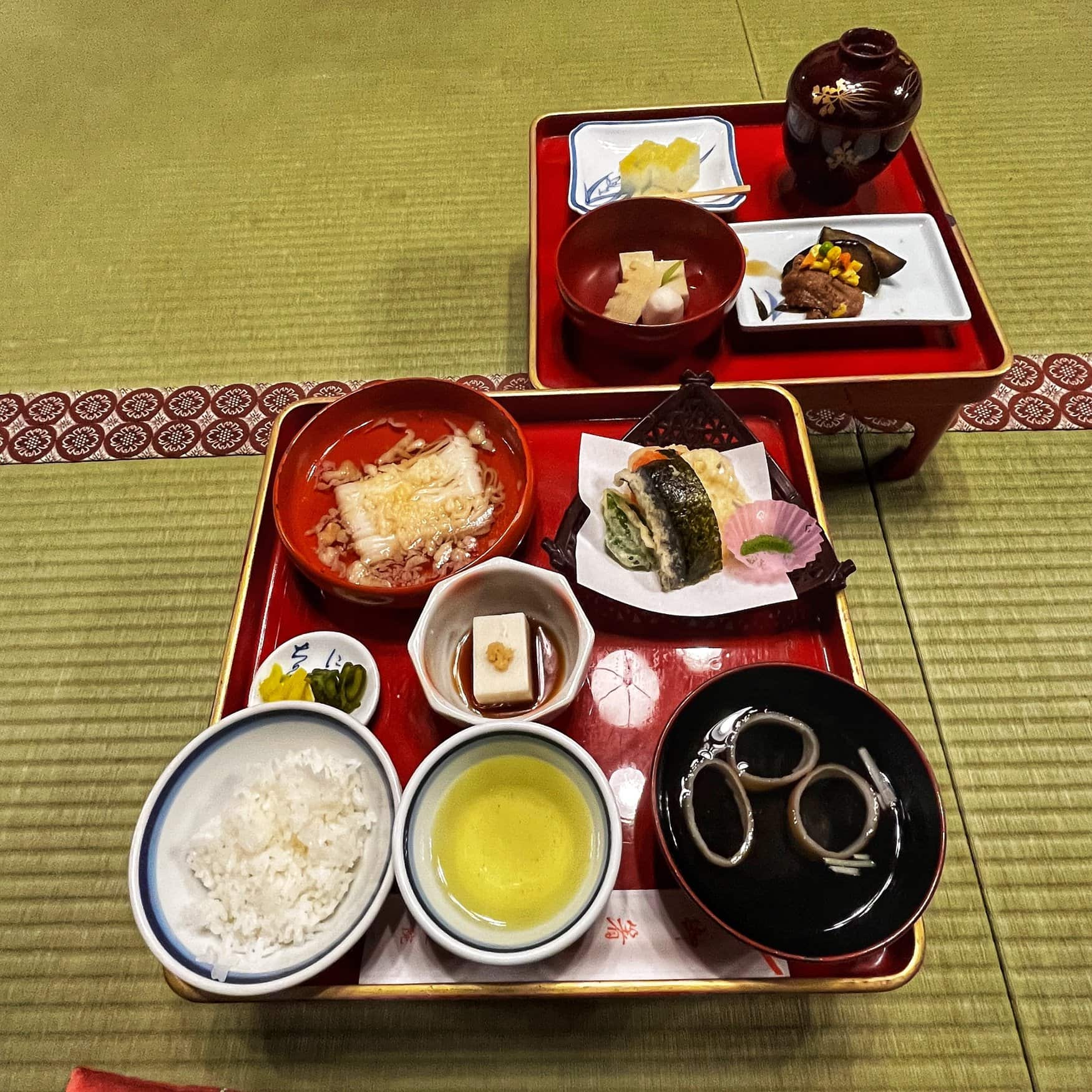 Traditionelle Tempelküche mit mehreren lackierten Tabletts und Schälchen auf Tatami, darauf Reis, Tofu, Gemüse und weitere Speisen.