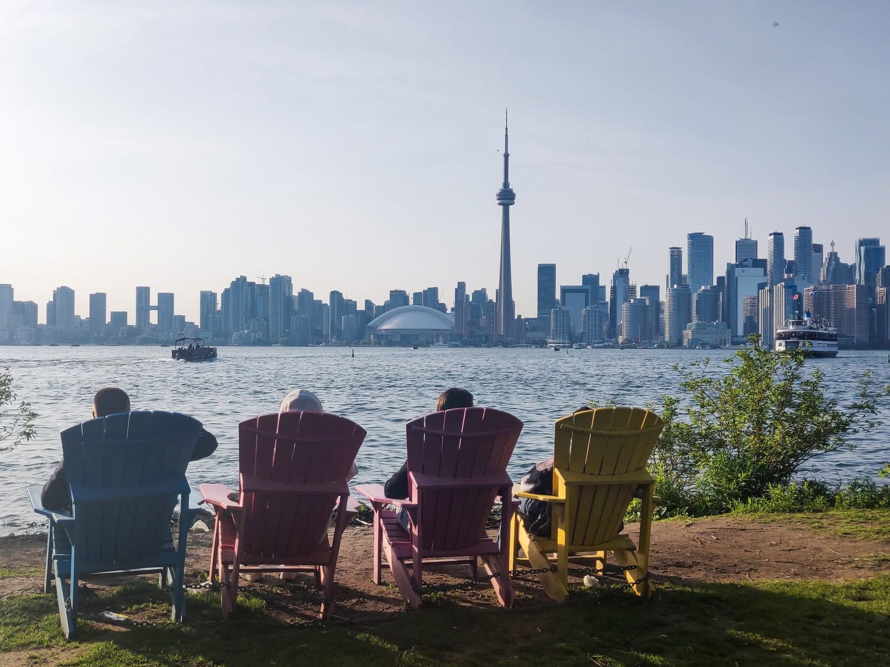 Blick vom Ufer des Lake Ontario auf die Skyline von Toronto. Am Ufer stehen vier bunte Liegestühle aus Holz, darauf vier Personen, die auf den See blicken