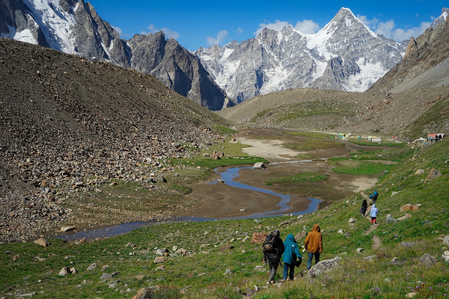 Wanderer steigen über einen Grashang zu einem Camp ab, dahinter vergletscherte Karakorum-Gipfel