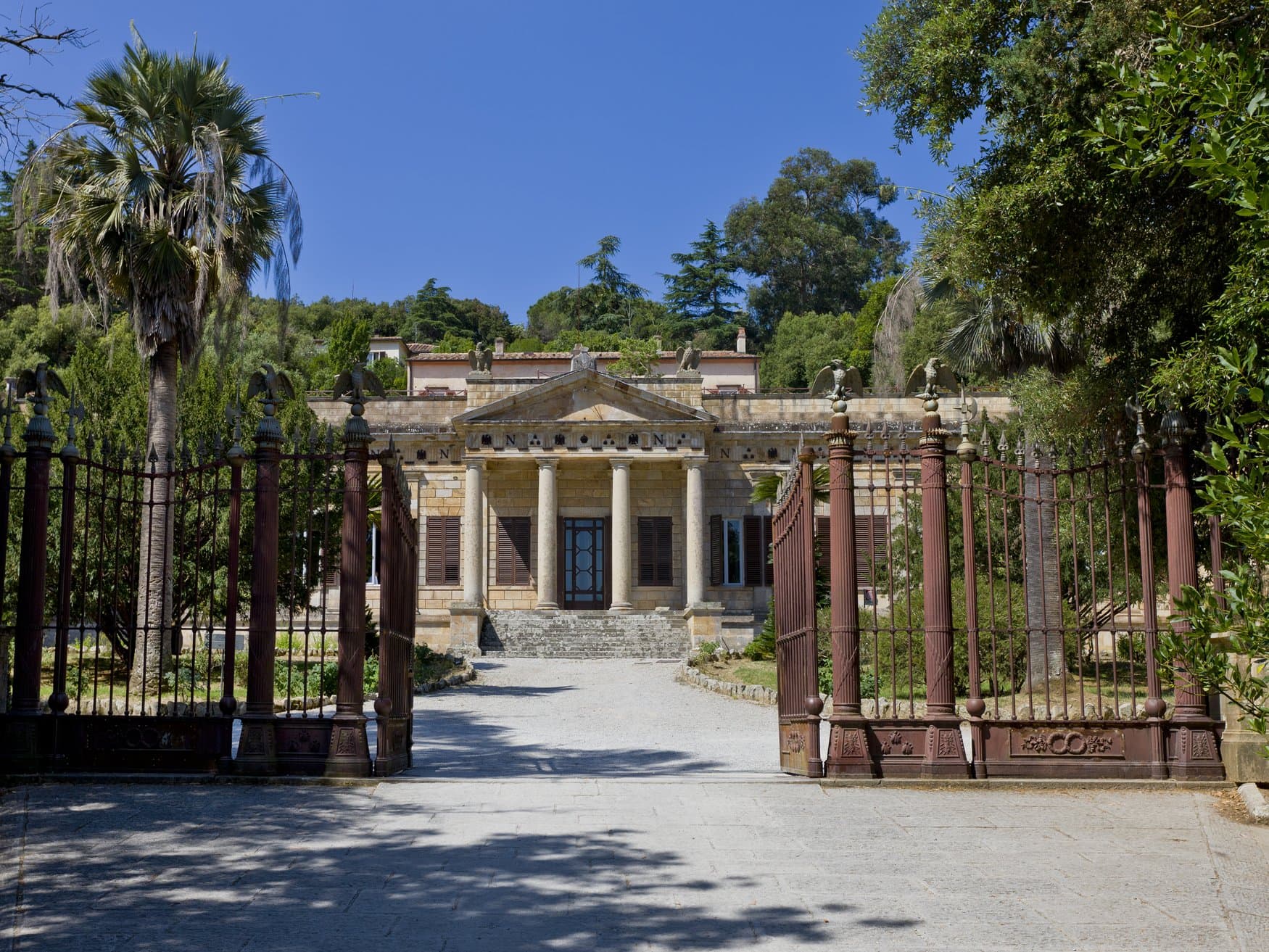 Blick durch das schmiedeeiserne Tor auf die Villa San Martino auf Elba, Napoleons Sommerresidenz. Die Fassade trägt einen Säulenportikus mit Giebel, auf den Torpfeilern sitzen Adlerfiguren.