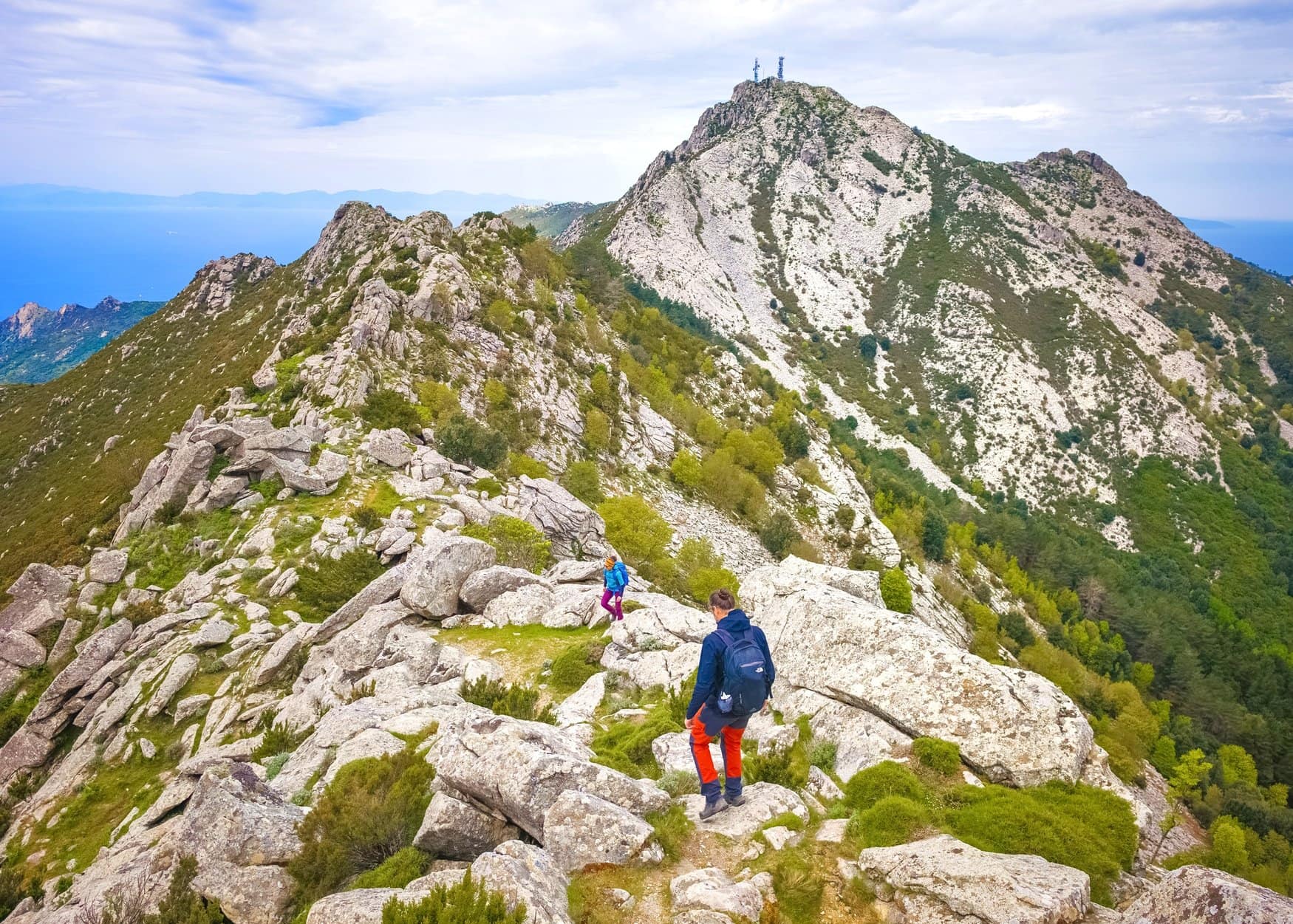 Zwei Wandernde gehen über einen felsigen Grat der Grande Traversata Elbana. Vor ihnen erhebt sich der Gipfel des Monte Capanne mit den Antennenmasten, links reicht der Blick aufs Meer.