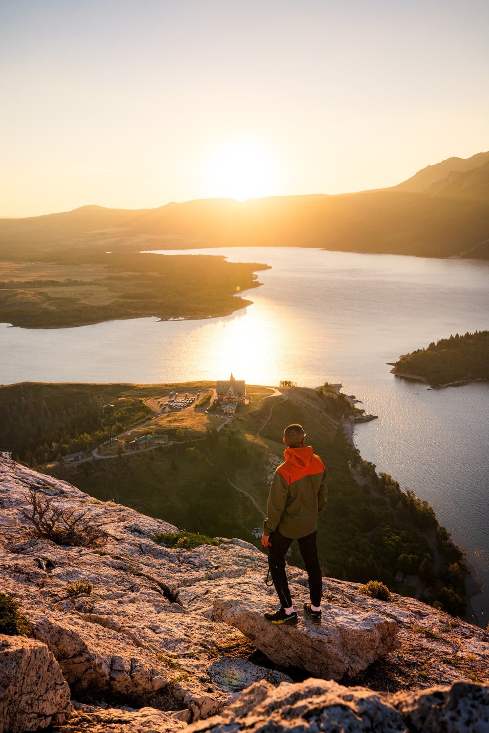 Wanderer in orangefarbener Jacke steht bei Sonnenaufgang auf dem Bear's Hump und blickt auf Waterton Township und den See tief unten