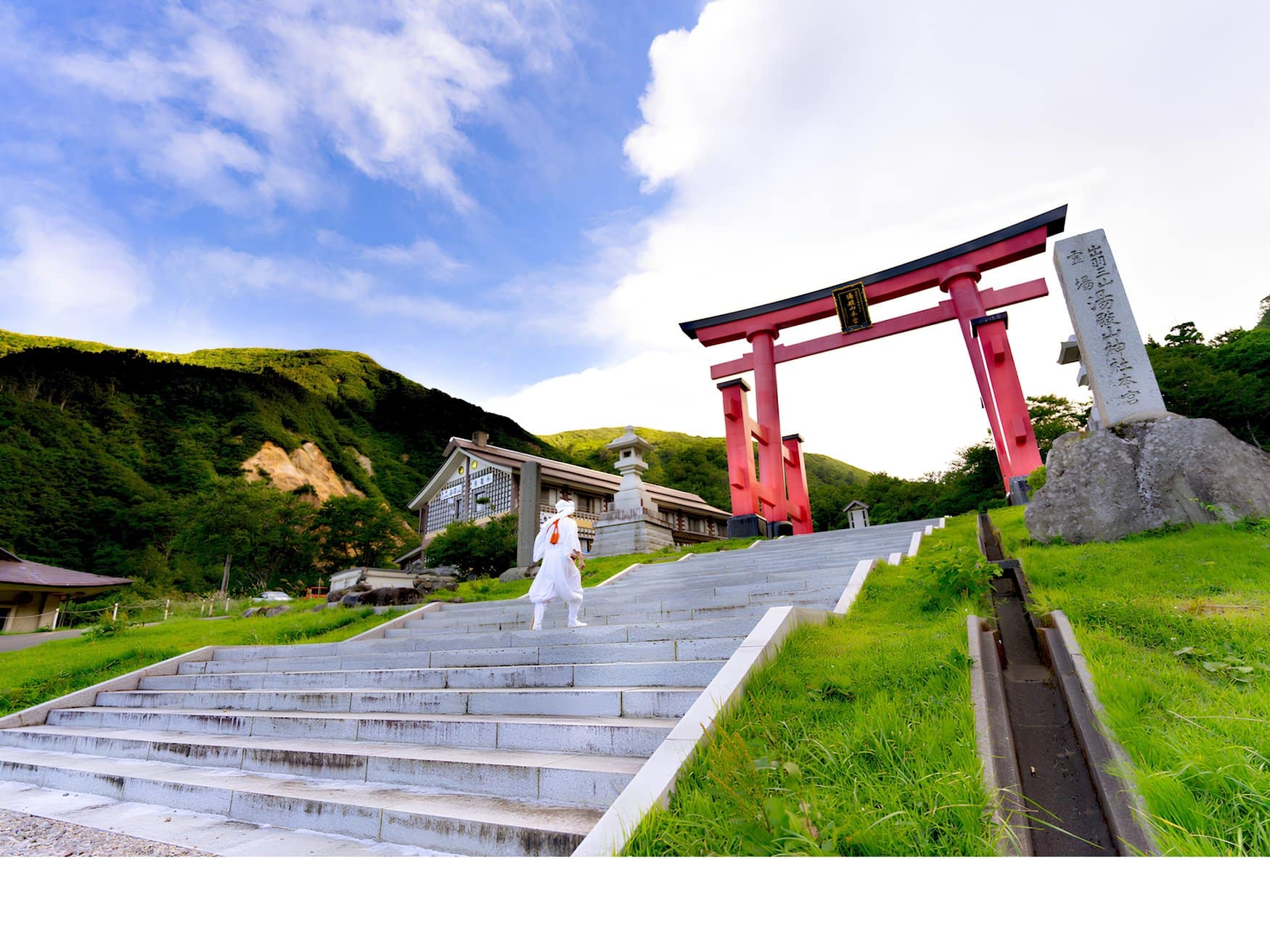 Eine Person in weißer Pilgerkleidung geht eine Steintreppe zu einem großen roten Torii hinauf, am Yudonosan im Dewa Sanzan.
