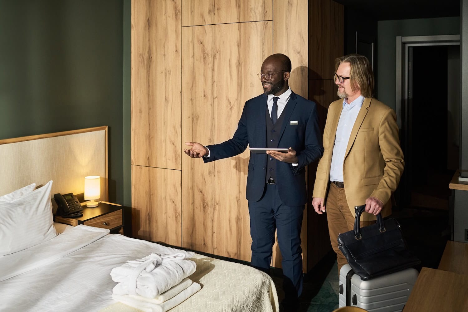 Hotelmanager begrüßt einen Gast in einem luxuriösen Hotelzimmer. Beide stehen neben dem Bett, auf dem ein geöffneter Koffer liegt. Der Manager hält ein Tablet in der Hand und lächelt freundlich. Die Szene vermittelt persönliche und aufmerksame Gastfreundschaft. Gefällt dir diese Persönlichkeit?