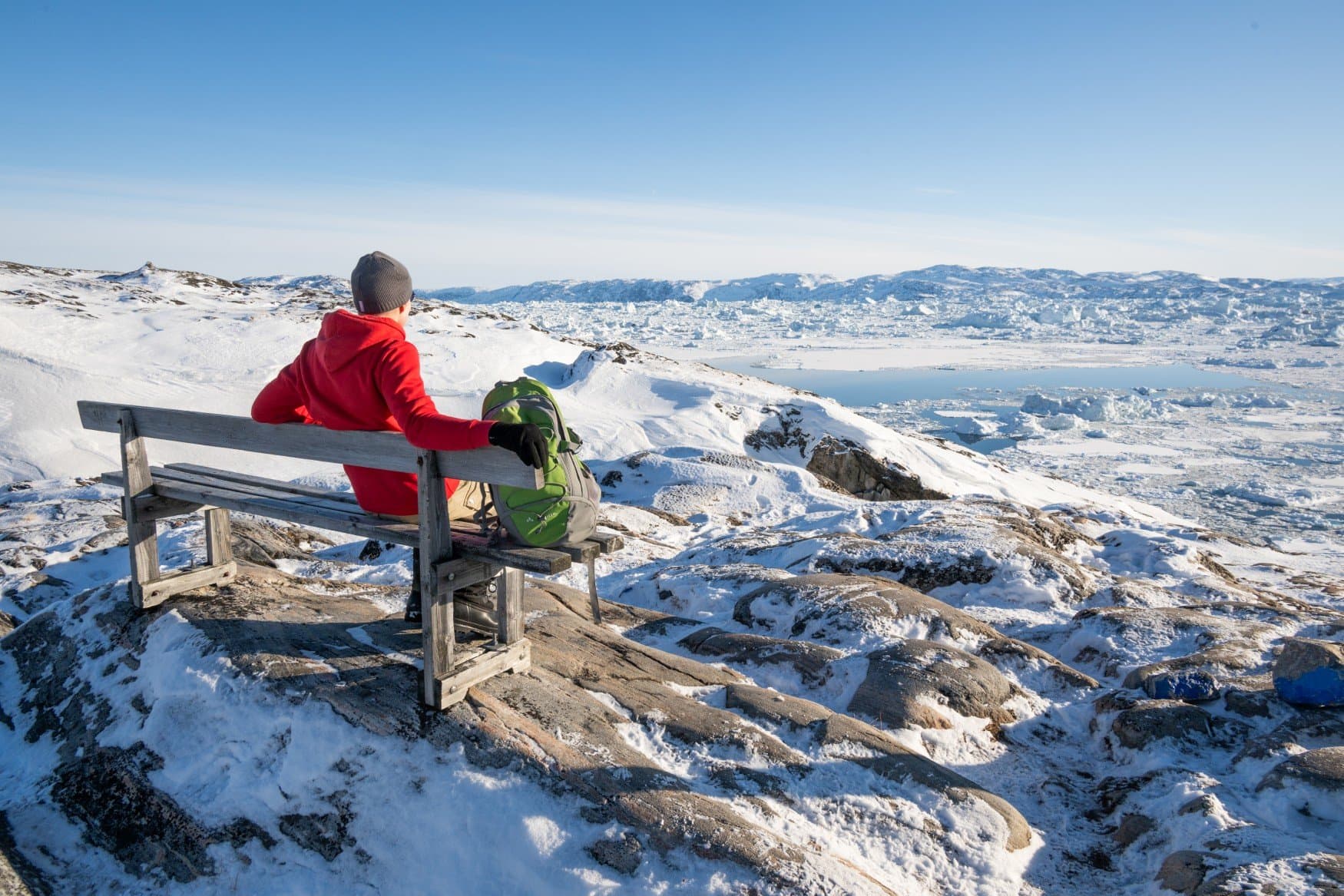 Eine Person in rotem Pullover sitzt mit Rucksack auf einer Holzbank auf einem Felsrücken und blickt über das Packeis des Eisfjords.