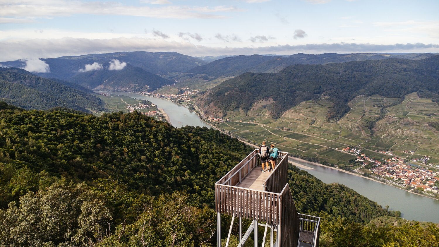 Zwei Wanderer stehen auf einer Aussichtsplattform hoch über der Wachau und blicken auf die Donau, umgeben von Weinterrassen und bewaldeten Hügeln.