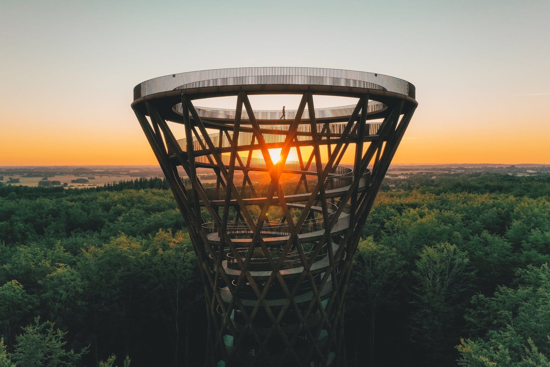 Aussichtsturm Skovtårnet in Dänemark, aufgenommen aus der Luft. Die trichterförmige Holzkonstruktion mit spiralförmiger Rampe steht über dem Wald, dahinter geht die Sonne über flachem Land auf. Auf der obersten Plattform steht eine Person.