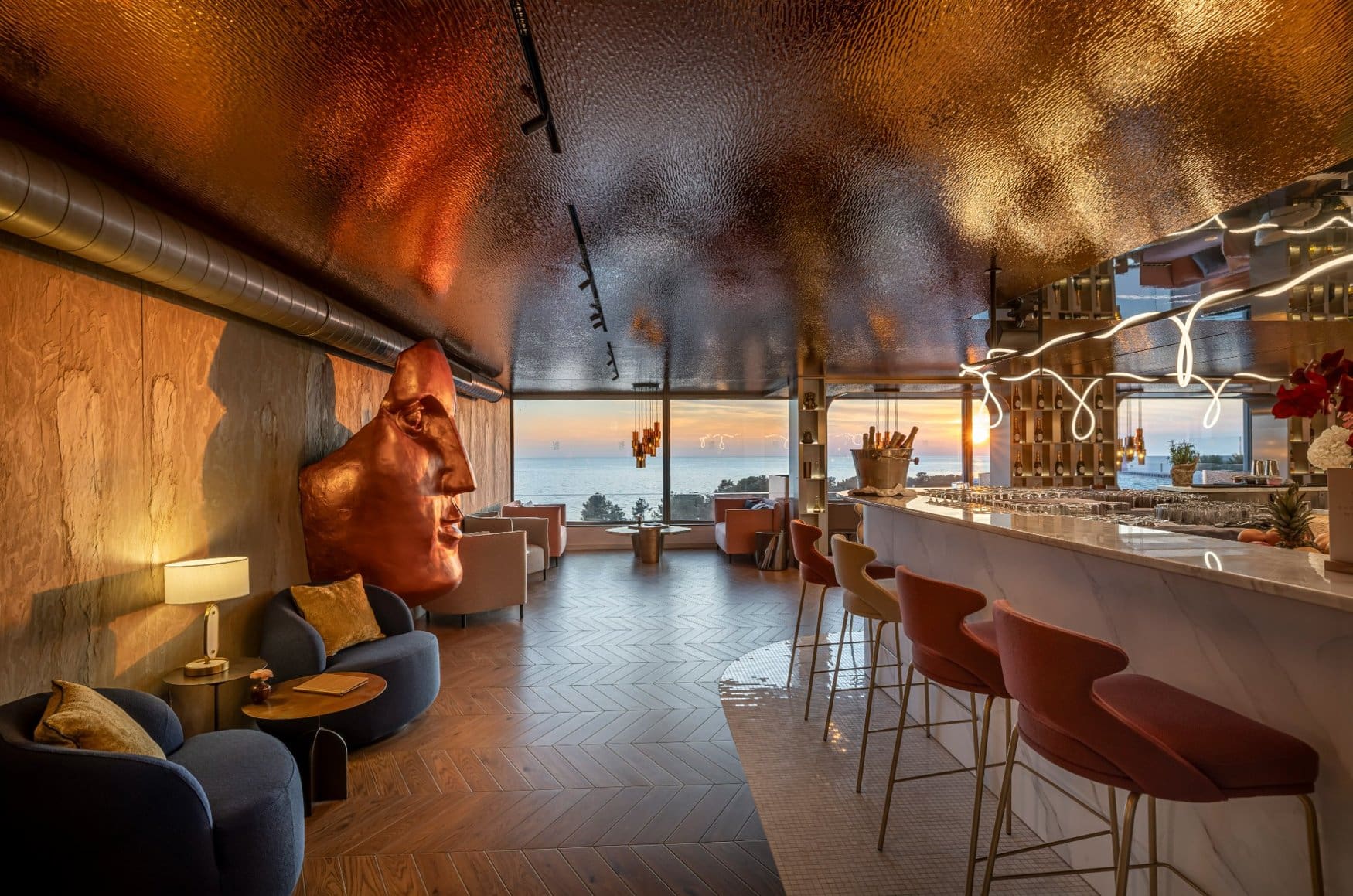 Eine Hotelbar mit gewölbter Decke aus kupferfarbenem Metall und einem Tresen aus hellem Marmor mit roten Barhockern. An der Wand steht eine große Kopfskulptur in Kupfer, durch die Fensterfront geht die Sonne über dem Meer unter.
