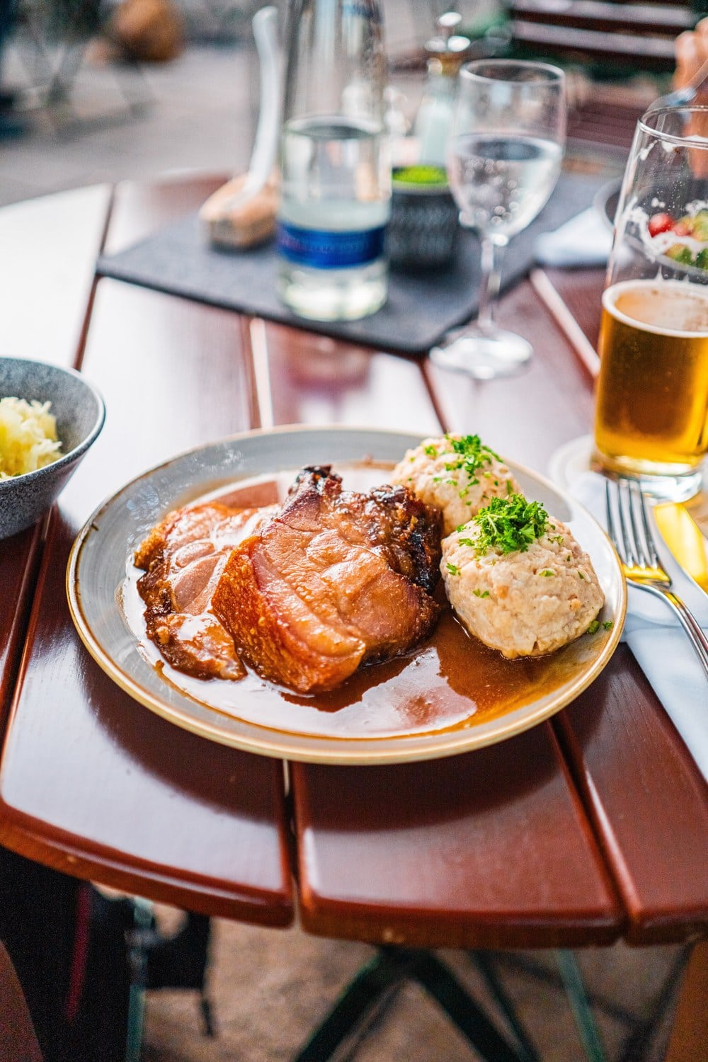Schweinebraten mit Kruste, zwei Semmelknödeln und dunkler Soße auf einem Holztisch im Freien, daneben ein Glas Bier und eine Schale Krautsalat.