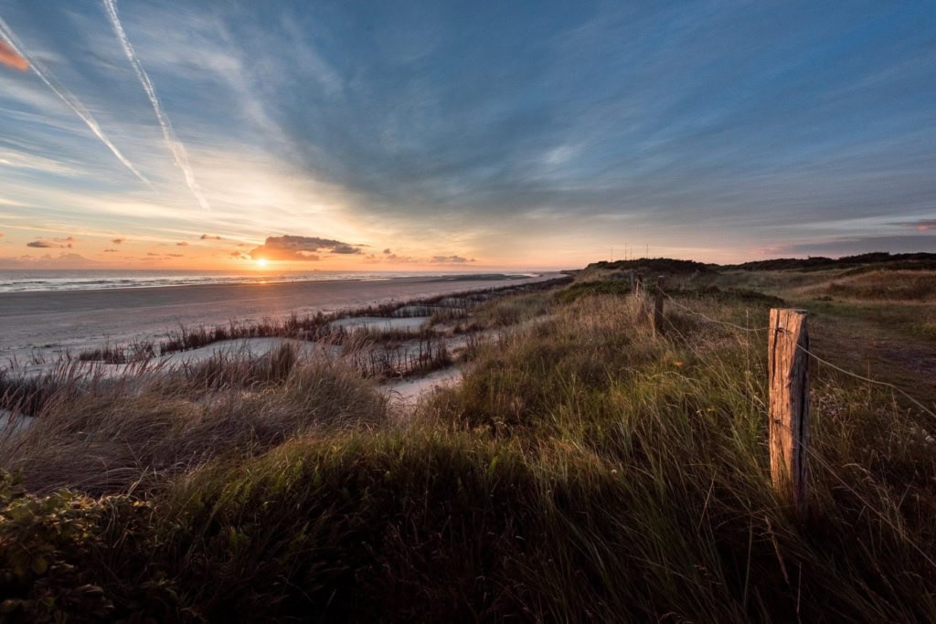 Blick von einer bewachsenen Düne im Osten von Wangerooge auf den breiten Strand. Im Vordergrund steht ein verwitterter Holzpfahl, über dem Meer steht die Sonne tief.
