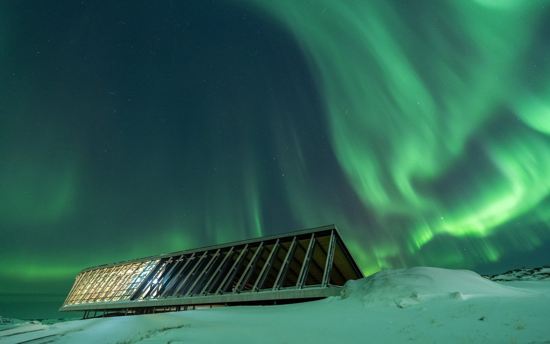 Ein langgestreckter Flachbau mit verdrehtem Dach und durchgehender Glasfassade liegt beleuchtet im Schnee, darüber zieht ein grünes Polarlicht in Schlieren über den Sternenhimmel.