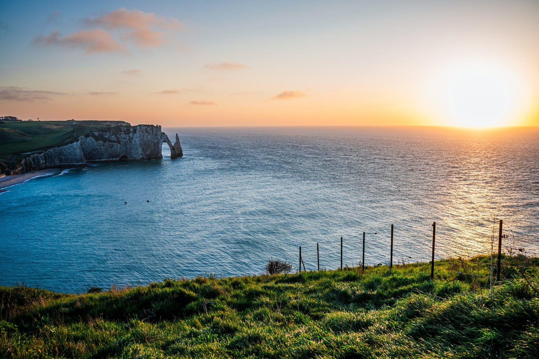 Die Kreidefelsen von Étretat in der Normandie bei Sonnenuntergang, mit dem Felsentor Porte d'Aval und der Nadel L'Aiguille. Im Vordergrund führt ein Zaun über eine Wiese zur Steilküste.