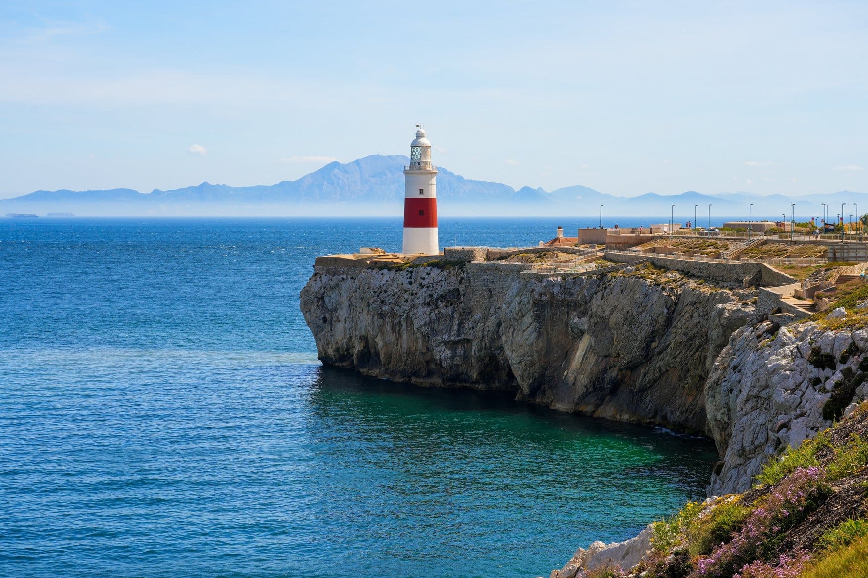 Der rot-weiß geringelte Leuchtturm am Europa Point in Gibraltar steht auf einer Kalksteinklippe über türkisfarbenem Wasser. Am Horizont erheben sich die Berge Marokkos.