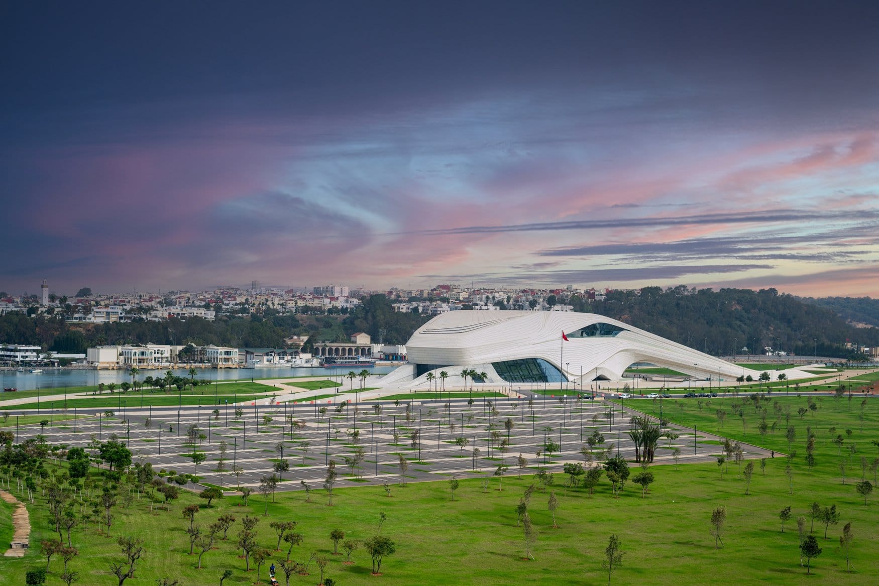 Das Grand Théâtre in Rabat am Ufer des Bouregreg in der Abenddämmerung. Der geschwungene weiße Baukörper liegt hinter Rasenflächen und einem Parkplatz, dahinter das Stadtbild von Salé.