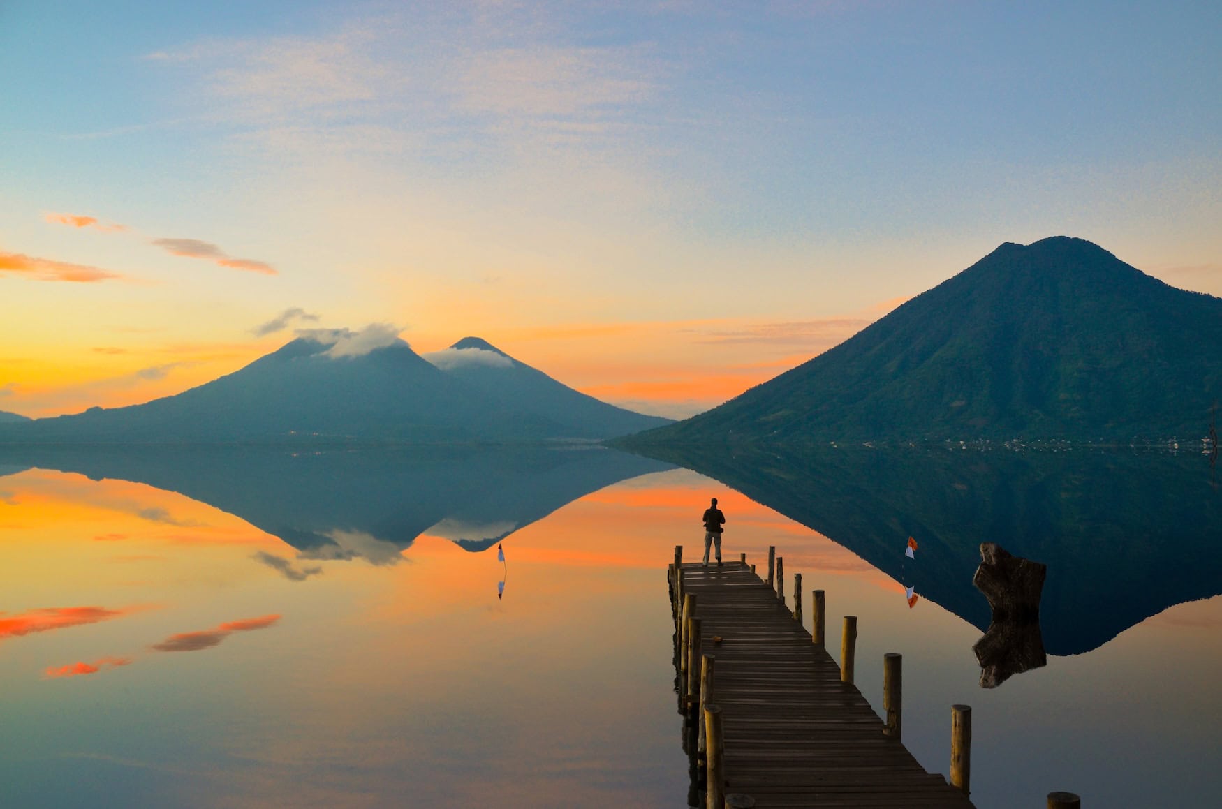 Sonnenaufgang am Atitlán-See in Guatemala, ein Holzsteg führt aufs Wasser, dahinter zwei Vulkane.