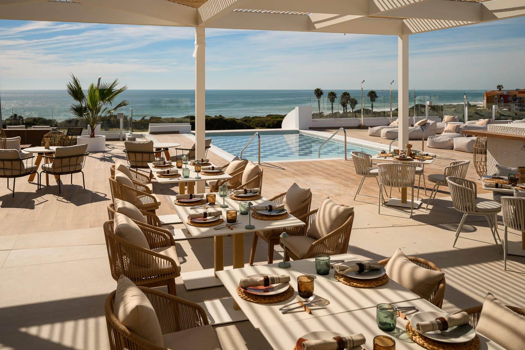 Die Terrasse des Restaurants Hayaca unter einer weißen Pergola, mit eingedeckten Tischen und Korbsesseln. Dahinter liegen ein Pool mit blauem Rautenmuster und Sitzsäcke, im Hintergrund die Brandung.