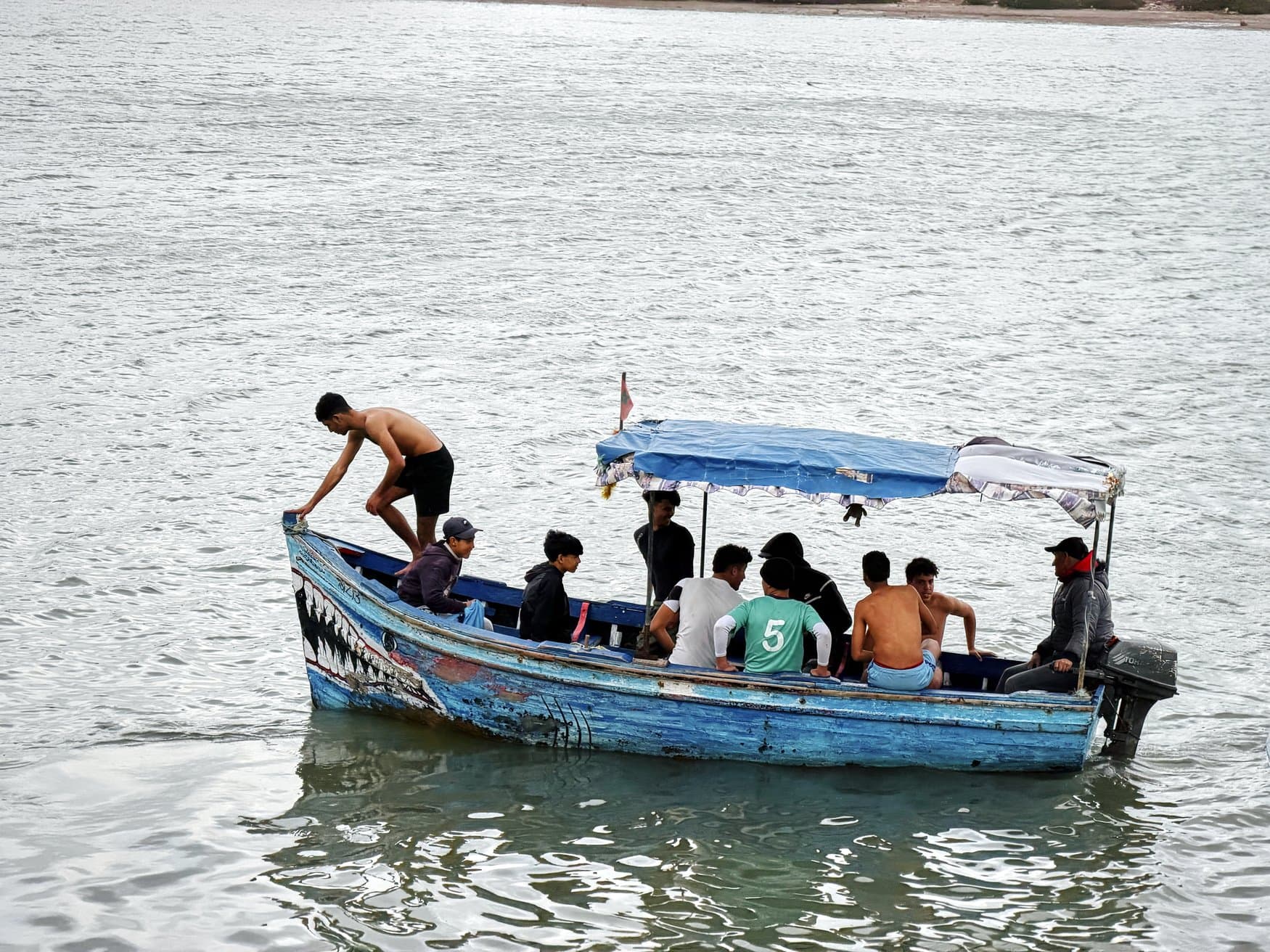 Ein blau gestrichenes Holzboot mit aufgemaltem Haifischmaul liegt auf ruhigem Wasser. Darin sitzen mehrere Jugendliche unter einem Sonnendach, am Heck steuert ein Mann den Außenborder.