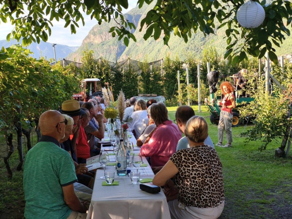 Gäste sitzen an einer langen weiß gedeckten Tafel zwischen Obstbäumen, im Hintergrund eine Sommeliere mit Weinflasche und ein Traktor. Über dem Tal steht eine steile Felswand.