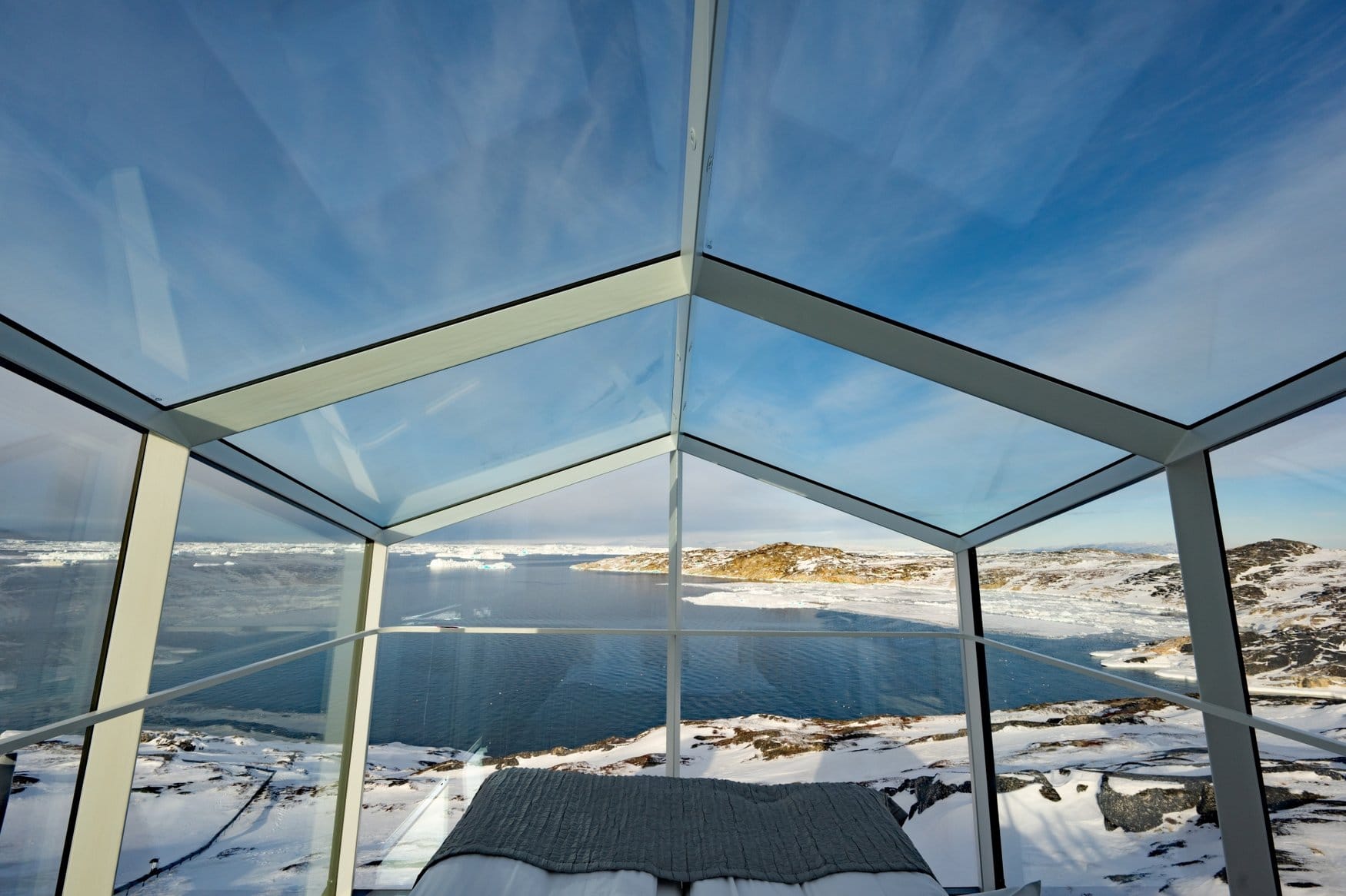 Blick aus einem verglasten Zimmer des Hotel Ilulissat über Dach und Wände hinweg auf den Fjord. Im Vordergrund liegt das Fußende eines Bettes mit grauer Decke, draußen treiben Eisschollen vor schneebedeckten Felshügeln.