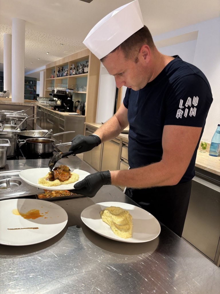 Ein Koch mit weißer Kochmütze und dunklem Laurin-Shirt richtet an der Buffetstation ein Fleischgericht auf Kartoffelpüree an.