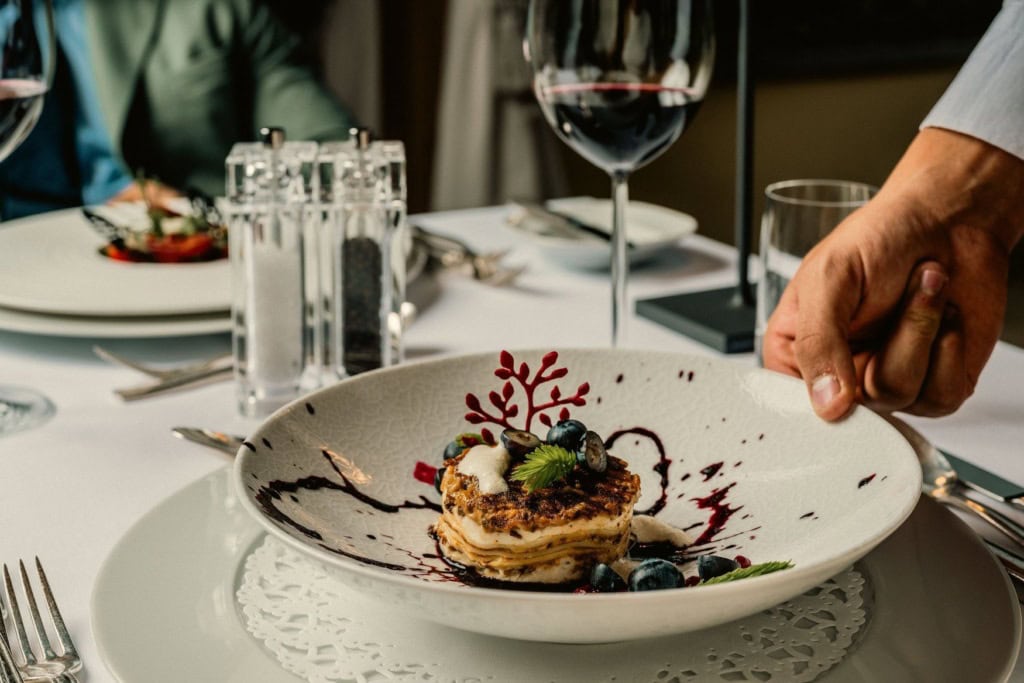 Eine Hand stellt einen tiefen Teller auf den gedeckten Tisch. Darauf ein geschichtetes Gericht mit gebräunter Oberfläche, Blaubeeren, Cremenocken und Fruchtsauce.