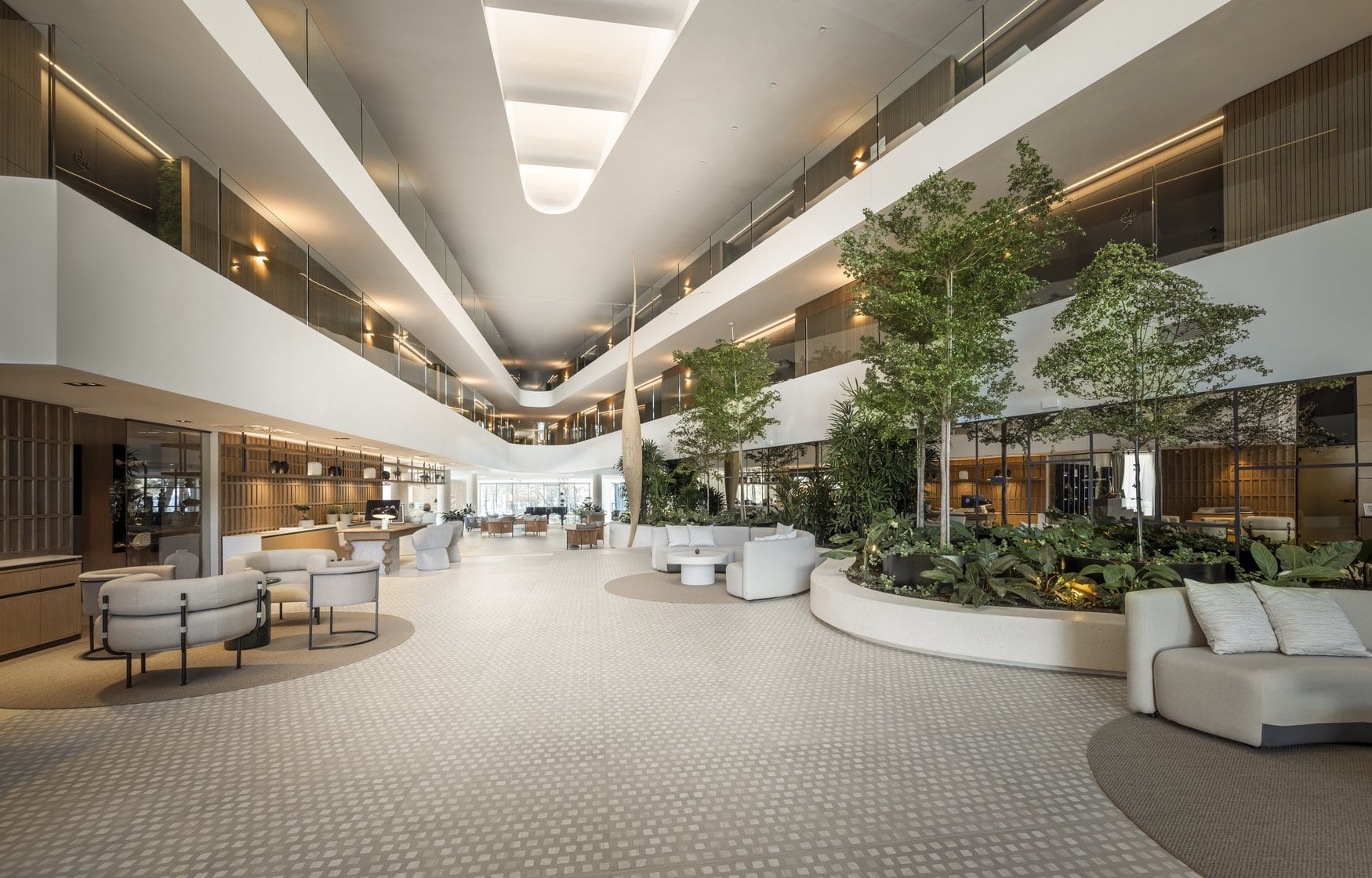 Die Lobby des Pical Resort über zwei Geschosse mit umlaufender Galerie. Links die Rezeption mit Holzregal, rechts ein bepflanztes Rundbeet mit Bäumen, davor helle Sitzgruppen auf gemustertem Boden.