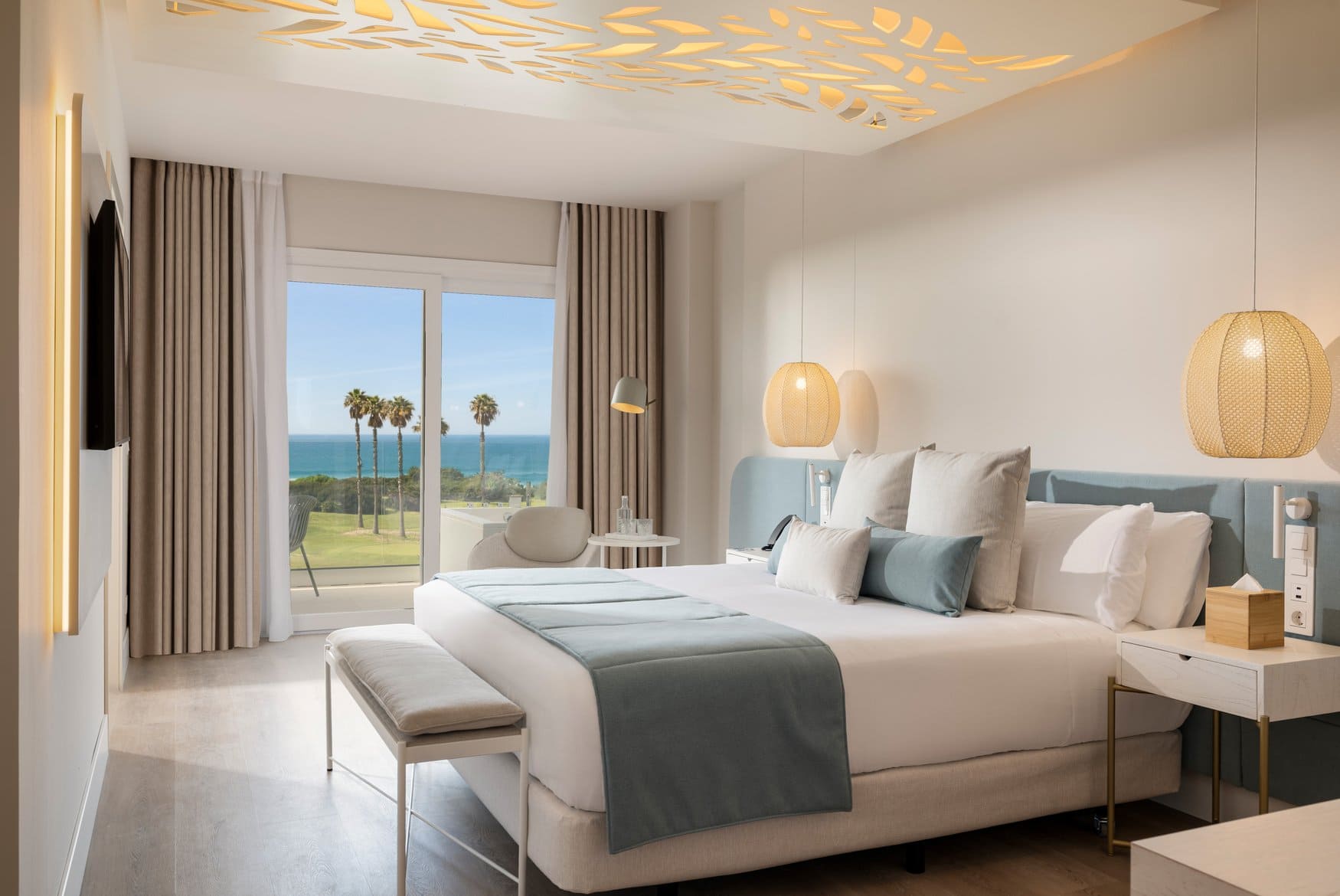 Das Zimmer Dolce Vita mit Doppelbett, hellblauem Kopfteil und einer Polsterbank am Fußende. Zwei Korbleuchten hängen von der Decke, durch die Balkontür sind Palmen und das Meer zu sehen.