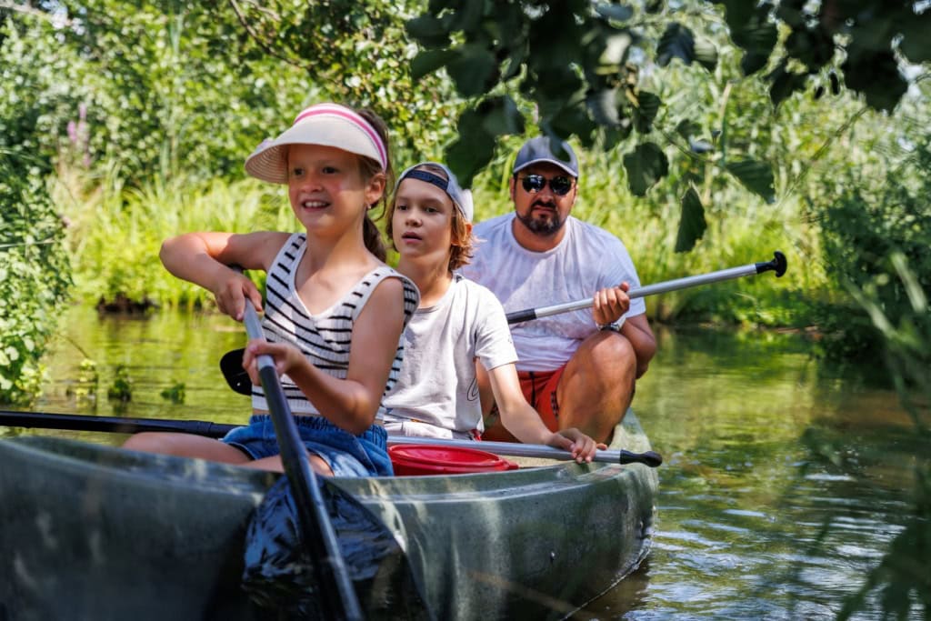 Ein Mann und zwei Kinder paddeln in einem Kanu über einen schmalen, von Grün gesäumten Fluss. Das Mädchen im Bug trägt ein Sonnenvisier, der Junge dahinter eine Basecap.