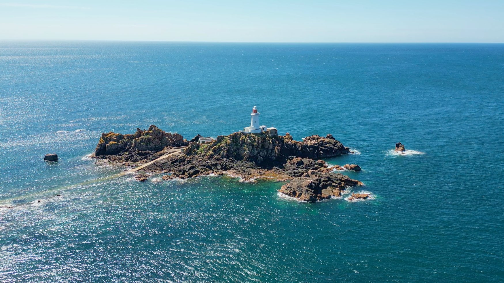Luftaufnahme des Leuchtturms La Corbière auf einer Felseninsel vor Jersey. Ein schmaler Damm verbindet die Insel mit dem Ufer, ringsum liegt türkisfarbenes Wasser.