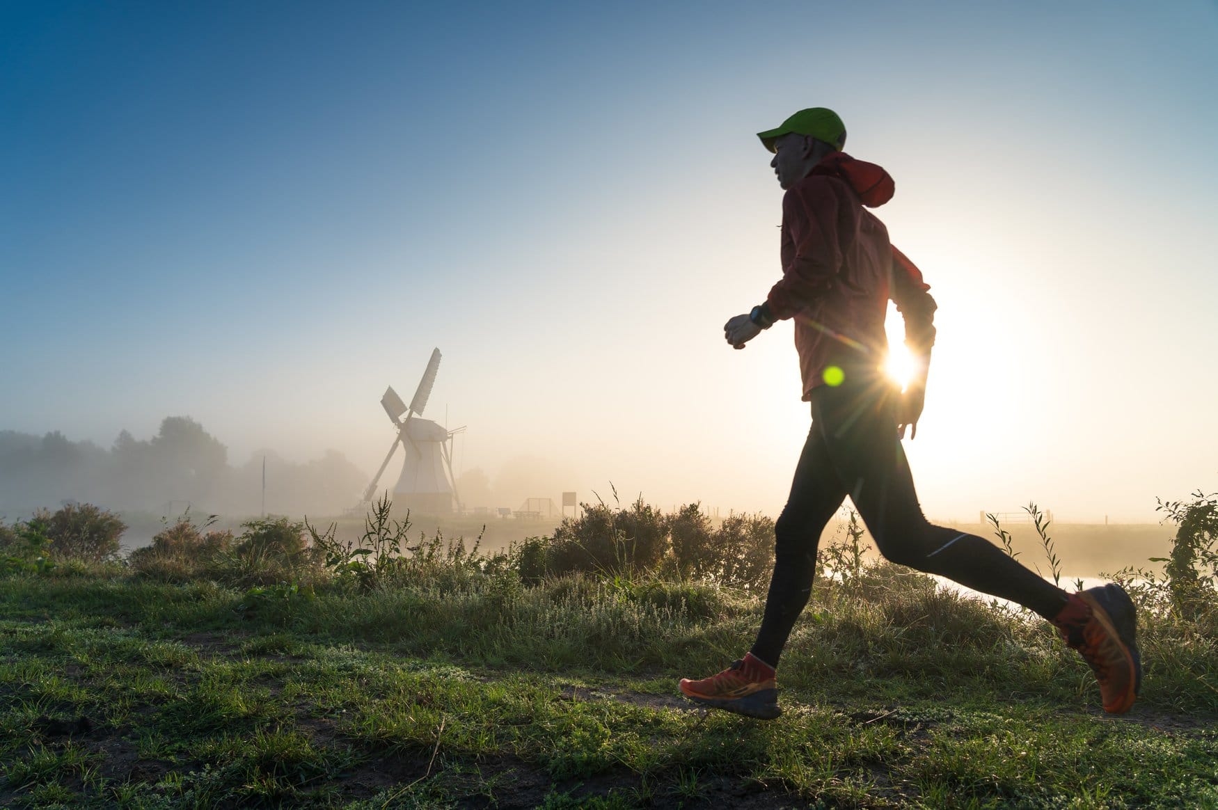 Ein Läufer im Gegenlicht auf einem Uferweg in den Niederlanden, im Nebel dahinter eine weiße Windmühle am Wasser.