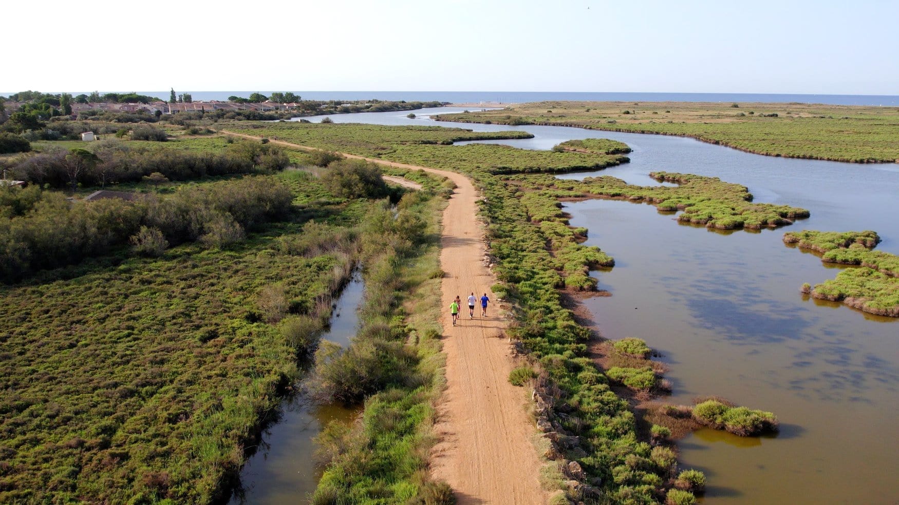 Luftaufnahme eines sandigen Damms zwischen flachen Lagunen, auf dem drei Personen laufen. Rechts liegt eine weite Wasserfläche mit Buschinseln, im Hintergrund eine Siedlung am Meer.